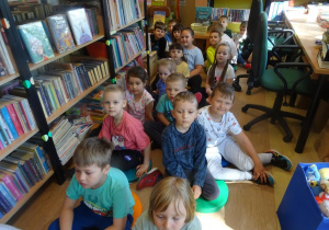 Dzieci siedzą pomiędzy regałami książek, słuchają wiersza.